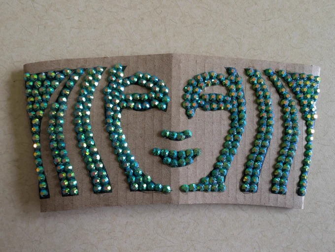 A standard brown cardboard Starbucks cup sleeve laid flat on a light textured surface. The sleeve features custom artwork depicting a stylized face meticulously formed out of small, shimmering, teal-and-green iridescent rhinestones. The nose and lips are outlined directly in the center, flanked by closed, serene eyes, while vertical curved lines of rhinestones sweep down both sides of the sleeve to frame the face.