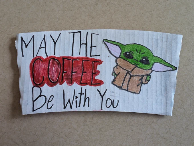 Hand-drawn artwork on a white corrugated Starbucks cup sleeve resting on a light textured surface. The left side features handwritten text in black marker that reads, "MAY THE COFFEE Be With You," with the word "COFFEE" emphasized in thick, stylized red bubble lettering. On the right side, there is a colored pencil illustration of Grogu (Baby Yoda) from Star Wars, with his signature large ears, green skin, and brown robes.