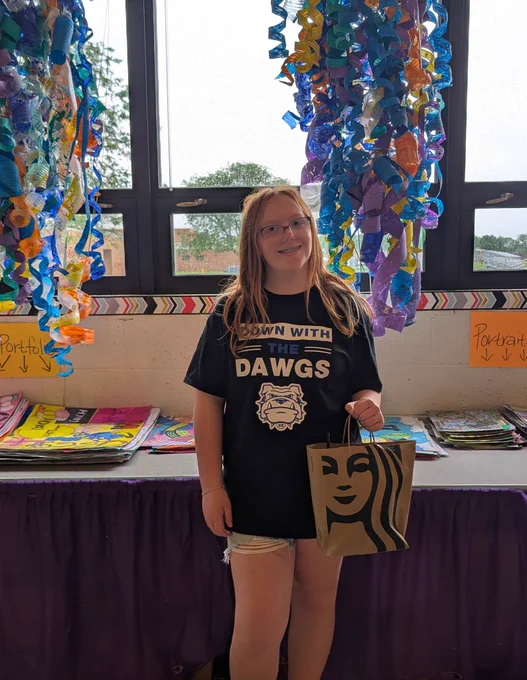 A full-length photo of a smiling young teenage girl with long reddish-brown hair and glasses, wearing a black T-shirt that reads "DOWN WITH THE DAWGS" above a white bulldog mascot graphic. She is holding a small brown paper Starbucks gift bag and standing in a classroom setting in front of large windows. Colorful, curled plastic art projects hang vertically on either side of her, and tables behind her are lined with stacks of student art portfolios and signs.