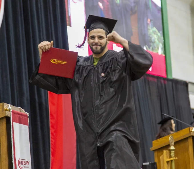 Graduate in black cap and gown proudly holding a red diploma cover on stage during commencement ceremony.