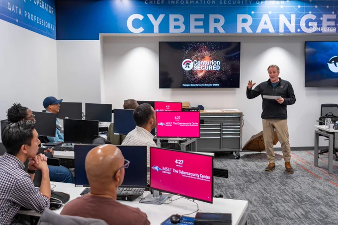 Instructor presenting cybersecurity concepts to a diverse group of adults in a modern training room labeled 'Cyber Range'.