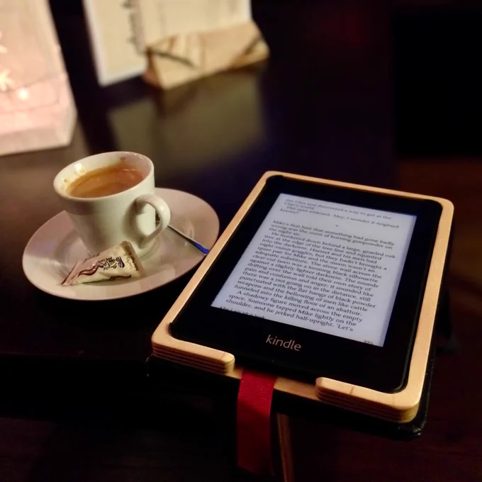 A Kindle e-reader resting on a table beside a small cup of espresso on a saucer, creating a cozy reading scene. The Kindle screen displays a page of text, suggesting someone relaxing with a book and coffee.
