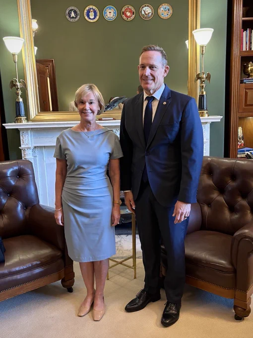 Ambassador and Senator standing and smiling in front of a living room set, looking off camera.