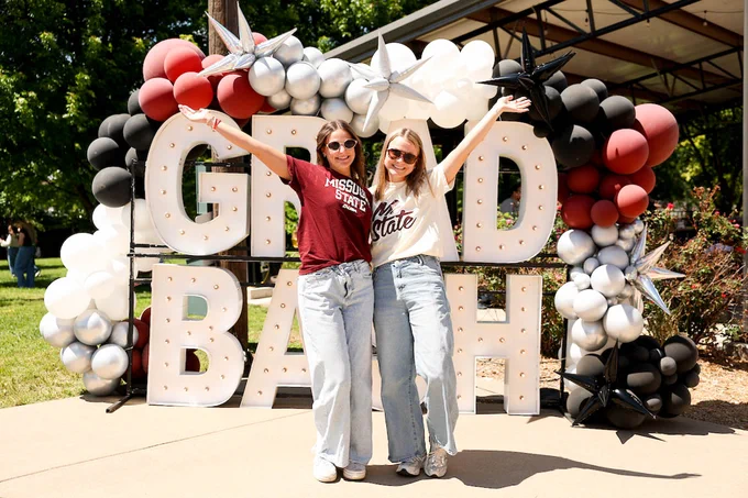 Two people stand smiling with arms raised in front of a large sign reading "GRAND BASH." The background features clusters of maroon, silver, black, and white balloons. Both individuals wear "Missouri State" T-shirts and sunglasses.