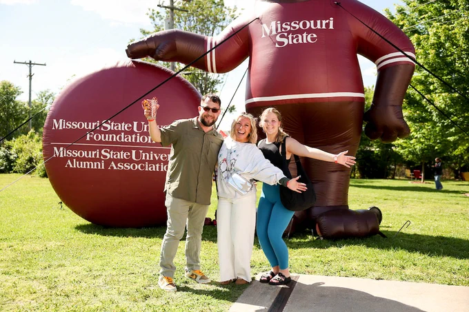 Three people are smiling and posing near large inflatable balloons displaying "Missouri State University Foundation" and "Alumni Association." Trees and grass are in the background.
