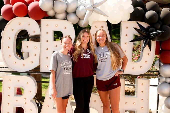 Three people are smiling and standing together in front of a "Grad Bash" sign. They are outdoors, surrounded by a balloon arch with red, black, gray, and white balloons. Two individuals are wearing shirts with "Missouri State" and one has "Bears" printed on it.