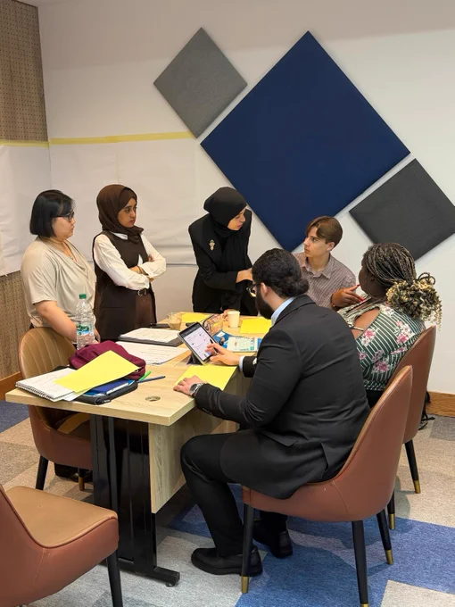 A group of young people surrounding a table, which has paper, pens, and other materials on it. Some participants are speaking to each other, while one participant scrolls on a tablet.