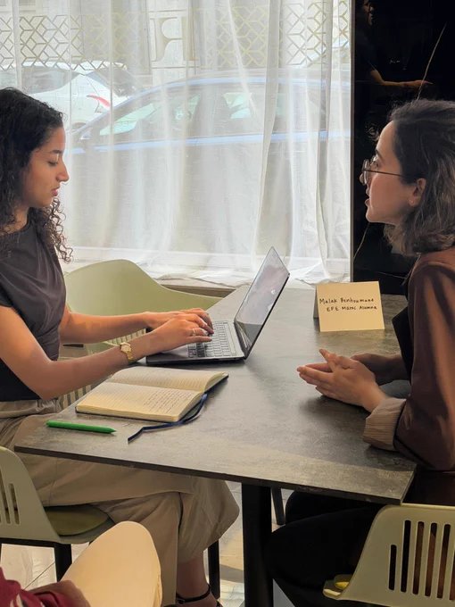 Two young people sitting at a table. One is on a laptop while the other is speaking.