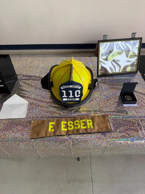 Picture of a yellow fire fighter helmet with the words "Wichita Airport 110 Esser" sitting on a table next to a retirement coin and a patch that says, "F. Esser."