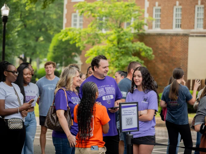 A group of people are gathered outdoors on a college campus, with some wearing shirts that say "University of Central Arkansas Dad." They appear to be participating in an event or gathering. A building with brickwork and windows is visible in the background, and there's a QR code on a sign in the foreground. Trees and greenery are on the left side of the image.
