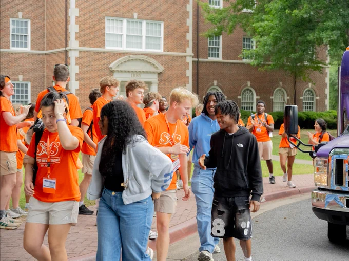 A group of people, mostly in orange shirts, are gathered near a building with arched windows. They appear to be in conversation and walking on a brick path. A purple Ford vehicle is parked nearby. Some individuals are wearing lanyards and name tags.