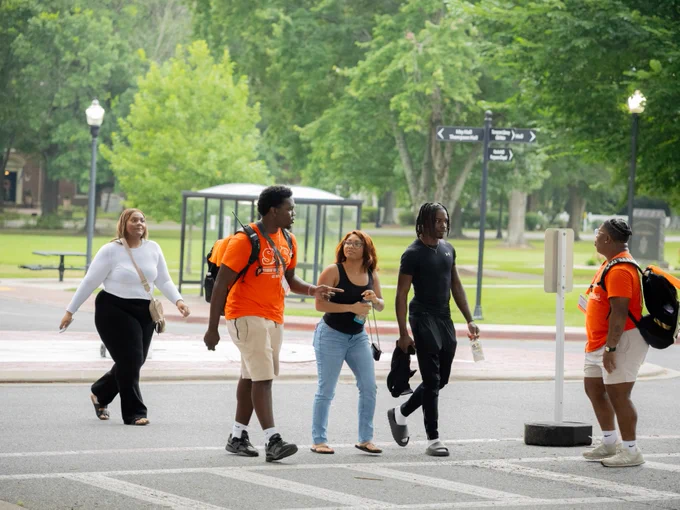A group of five people walking across a street on a leafy campus. Two individuals wear bright orange shirts. In the background, there are green trees, a bus shelter, and directional signs.