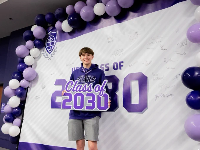 A person is standing in front of a banner that reads "Class of 2030" with the word "Bears" on their shirt. The banner is decorated with a border of purple and white balloons. The wall is signed by various individuals.