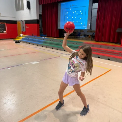 A young girl wearing a colorful shirt and holding a red ball, standing on a gymnasium floor with a screen and curtain visible in the background.