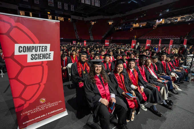 A group of graduates seated on the Xfinity Center floor, with a red Computer Science banner on the left