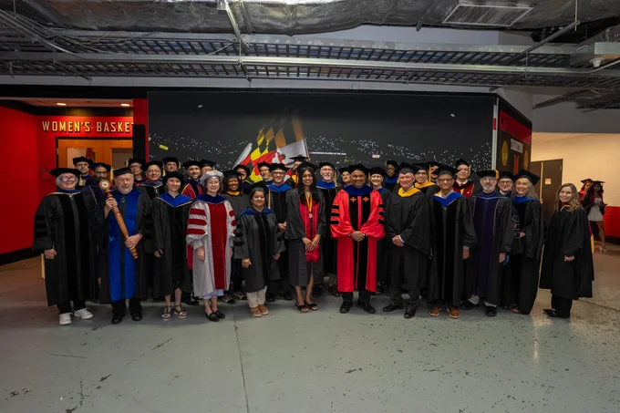 A group of more than 40 people in graduation regalia standing in front of a mural in a hallway 