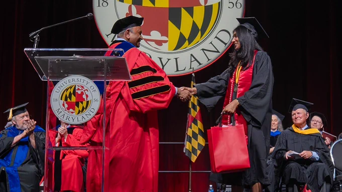 Dean Amitabh Varshney shaking a graduate's hand. The graduate is holding a red giftbag. A group of people behind them is clapping and smiling.
