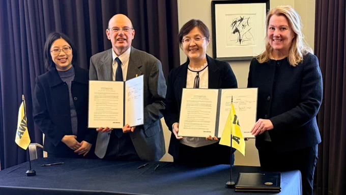 Four people stand behind a table during a formal signing event, smiling toward the camera. Two individuals hold open signed documents in presentation folders. Small University of Iowa flags sit on the table in front of them, with dark curtains and framed artwork in the background.