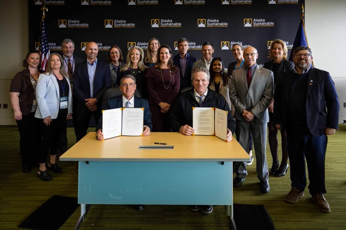 A group of individuals is posed in a professional setting. Two people are seated at a table in the foreground, holding documents. Behind them, others stand in front of a backdrop displaying the Alaska Sustainable logo.