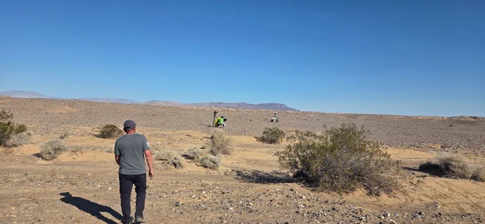 Man walking through a dry, rocky desert landscape with sparse bushes under a clear blue sky.