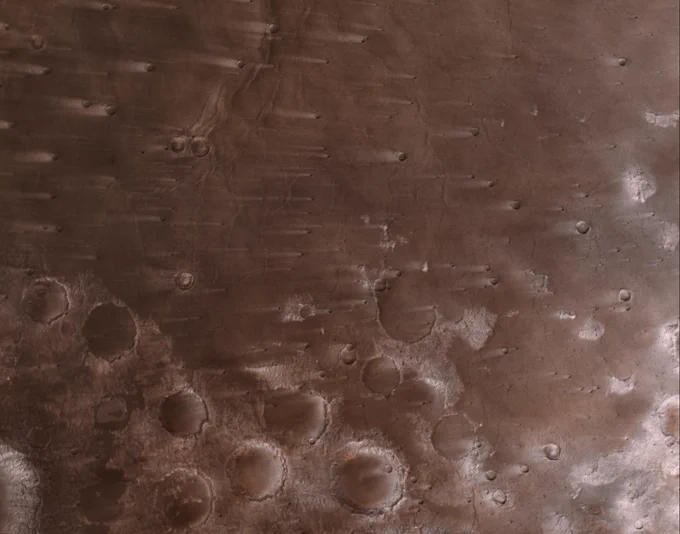 A bird\u2019s-eye view of the rusty-red Martian surface. The surface is heavily cratered with white outlines around them, streaky due to the planet\u2019s wind. Credit: NASA/JPL-Caltech/ASU