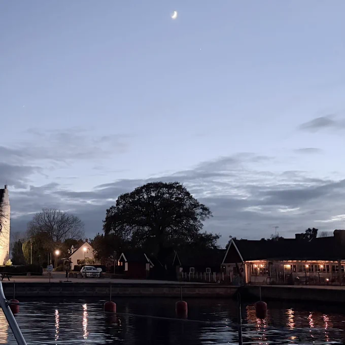 Abendhimmel über dem Hafen von Kristianopel in Blekinge, Schweden. Im Vordergrund eine Wasserfläche, dunkelblau mit sanften Wellen, in der Bildmitte Häuser und karg belaubte Bäume darüber wo hellblauer Himmel mit ein paar Wolken. Ganz oben am Bildrand die Mondsichel.
Ein Bild, das viel Ruhe transportiert.