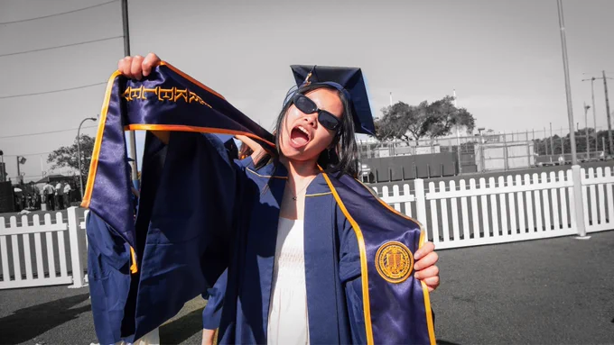A UC Berkeley graduate in blue-and-gold regalia smiles widely while holding up their stole, with the background in black and white.