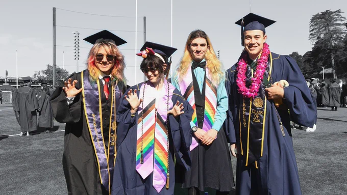 Four UC Berkeley graduates pose on the field in caps, gowns and colorful stoles, with the background mostly in black and white.