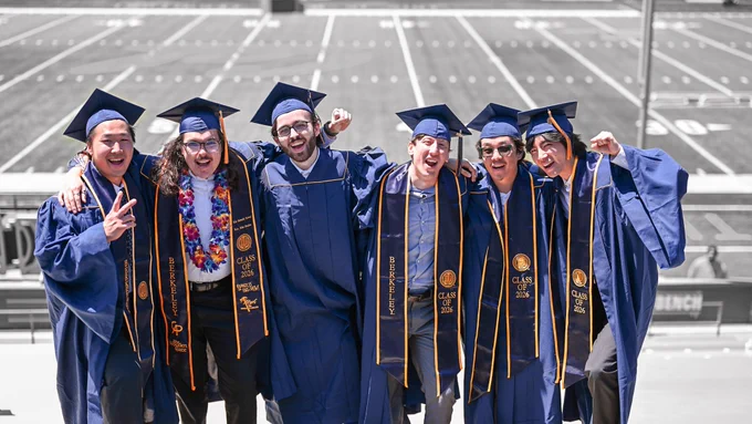 Six UC Berkeley graduates in blue-and-gold commencement regalia pose together with arms around each other inside the stadium.