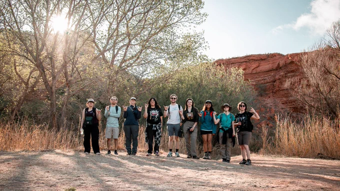 A group of people standing outdoors in a sunlit area with trees and a red rocky cliff in the background. They are casually dressed and appear to be enjoying a hike, with some smiling and waving at the camera.