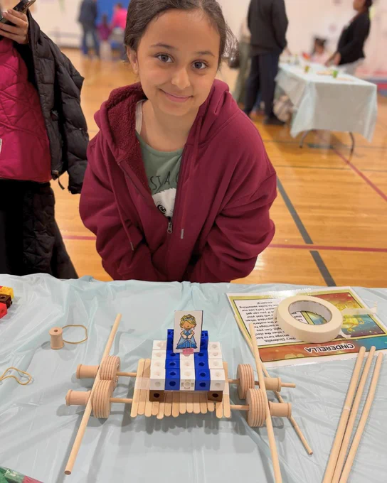 Child in a maroon hoodie sitting behind a table displaying a wooden and plastic block vehicle model at an indoor event.