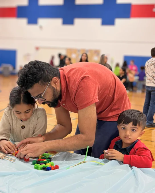 Man in a red shirt helping two children build with colorful blocks on a table in a gymnasium.