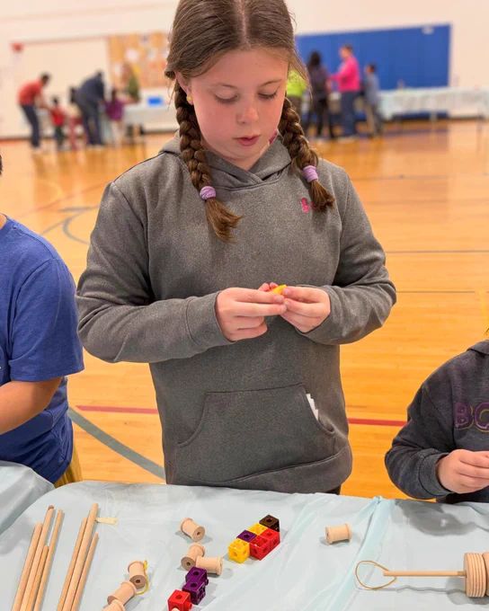Child with braided hair wearing a gray hoodie assembling colorful plastic blocks at a table in a gymnasium setting.