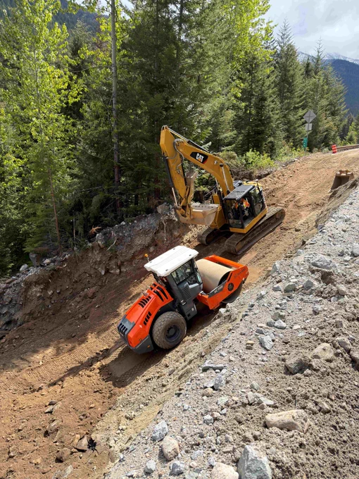 An orange compactor and yellow excavator rebuild an embankment along a washed out section of State Route 20 in a rural area.
