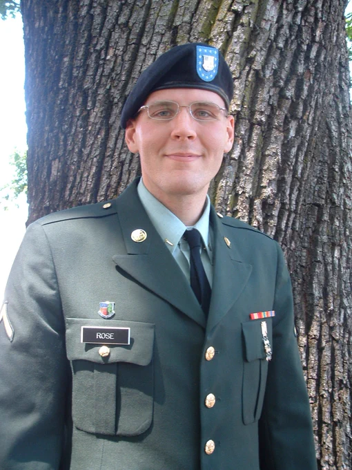 A person in a military uniform stands in front of a large tree. The uniform has a name tag that reads "Rose" and features various insignia and medals. The person wears a beret as part of the attire.