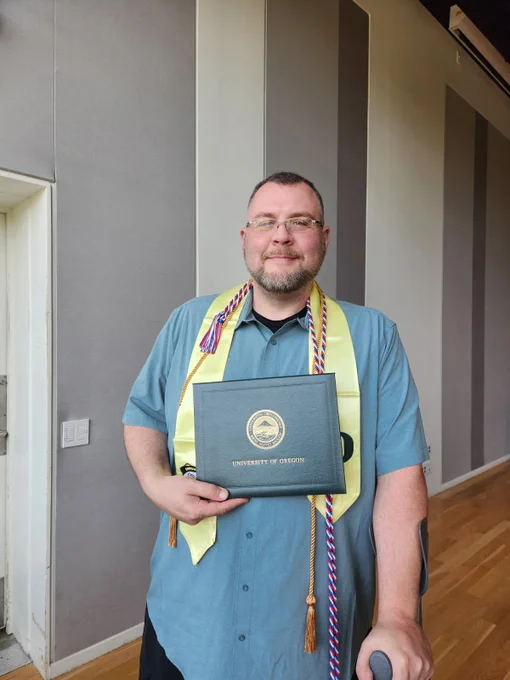 A person wearing a blue shirt is holding a University of Oregon certificate and is adorned with a yellow stole and honor cords. They are standing indoors on a wooden floor, with striped wall panels in the background.