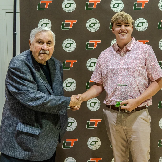 Rod Harmon shakes hands with Grayson Pinkston as Pinkston holds the J. Rod Harmon Excellence in Service Award during Lincoln Trail College Commencement. They stand in front of an LTC backdrop featuring the college logo.