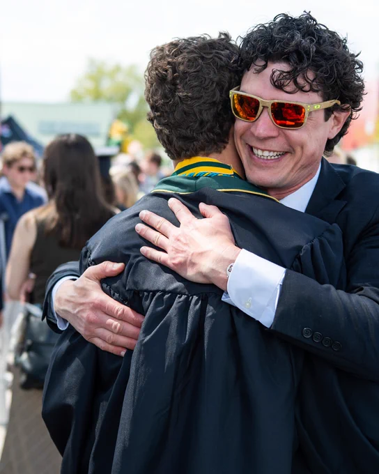 Two people embracing at a graduation ceremony. One is wearing a cap and gown, while the other is in a suit and wearing reflective sunglasses. The background shows other attendees and greenery.