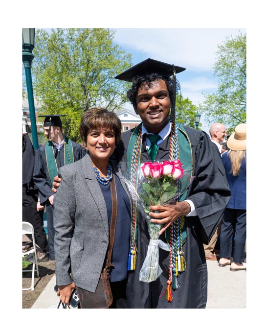 A person in a graduation cap and gown holds a bouquet of roses, standing beside another person who is smiling. They are outdoors, with trees and people in the background.
