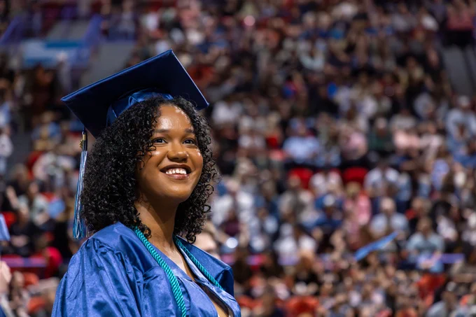 A graduate in a blue cap and gown smiles in a crowded auditorium filled with spectators.