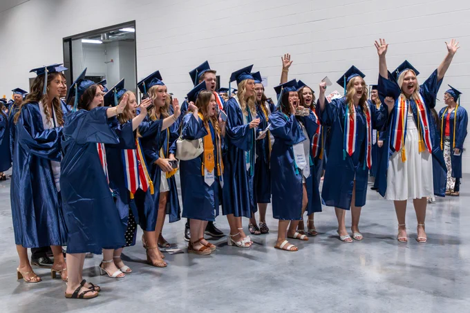 A group of people wearing blue graduation caps and gowns celebrate enthusiastically.