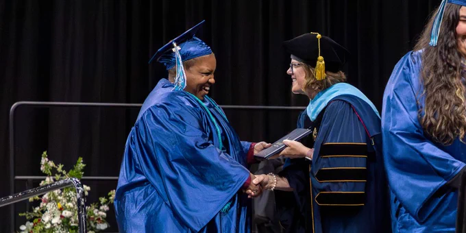 A graduate in a blue cap and gown is shaking hands with a person in academic regalia on stage during a graduation ceremony. 