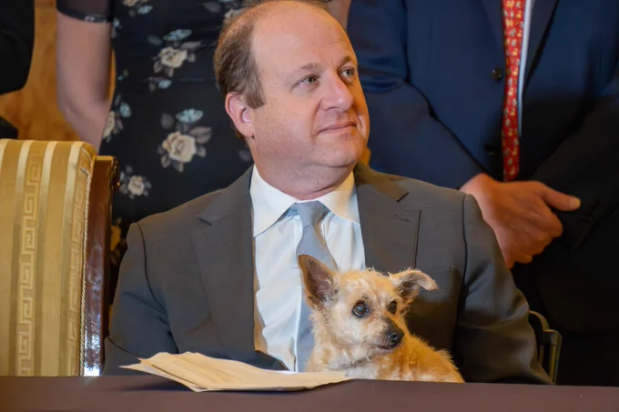 Small tan dog sitting calmly on Governor Polis’s lap during a formal event, with several people standing behind them.