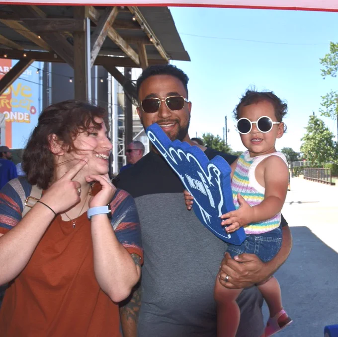 A family of three stands outdoors at Karbach Brewing Co. during the TAMU-CC Islander Alumni Association Houston Chapter Mixer. A TAMU-CC alumnus holds a large blue foam hand. A child is wearing sunglasses and sits in their arms. There are trees and a building in the background.