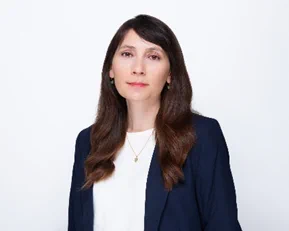 Professional headshot of a woman with long, dark brown hair wearing a navy blazer over a white top, facing forward against a light gray background.