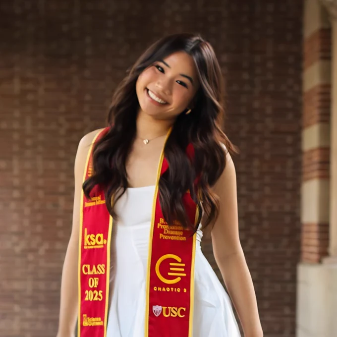 A person wearing a white outfit and a red graduation stole with yellow trim. The stole features the text "Class of 2025," "USC," and other insignia. Brick wall background.