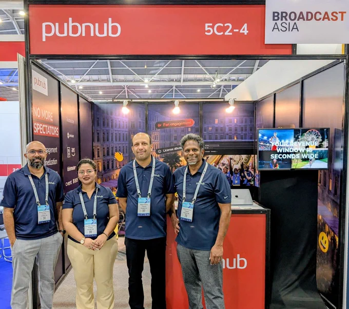 A group of four people poses in front of the PubNub booth at a trade show. The booth is part of the Broadcast Asia event, featuring promotional text and technology displays.