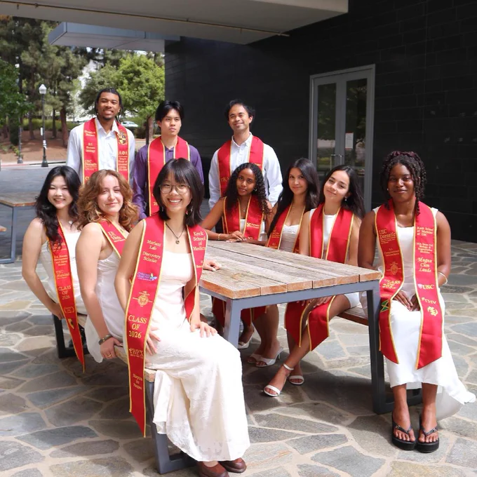 A group of people wearing white outfits and red stoles sit and stand around a wooden bench outdoors. They appear to be celebrating a graduation event. Trees and a building are visible in the background.