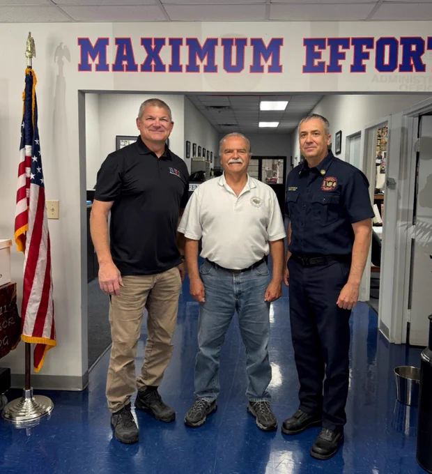 Three people are standing in a hallway with blue flooring, beneath a sign that reads "MAXIMUM EFFORT." An American flag is positioned to their left. They appear to be in a professional setting.