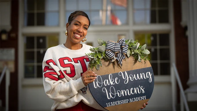 Dr. Mariah Walker wearing an NC State sweatshirt and holding a round wooden sign decorated with greenery and a striped bow that reads 'Wake Young Women's Leadership Academy'.